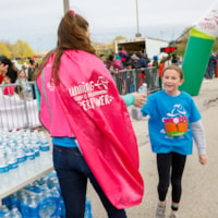 Volunteer wearing a "Running is my Superpower" cape hands water to a GOTR girl at the Finish Line Chute