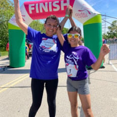 A Girls on the Run participant and 5K buddy raise their arms together in celebration under the 5K Finish Arch.